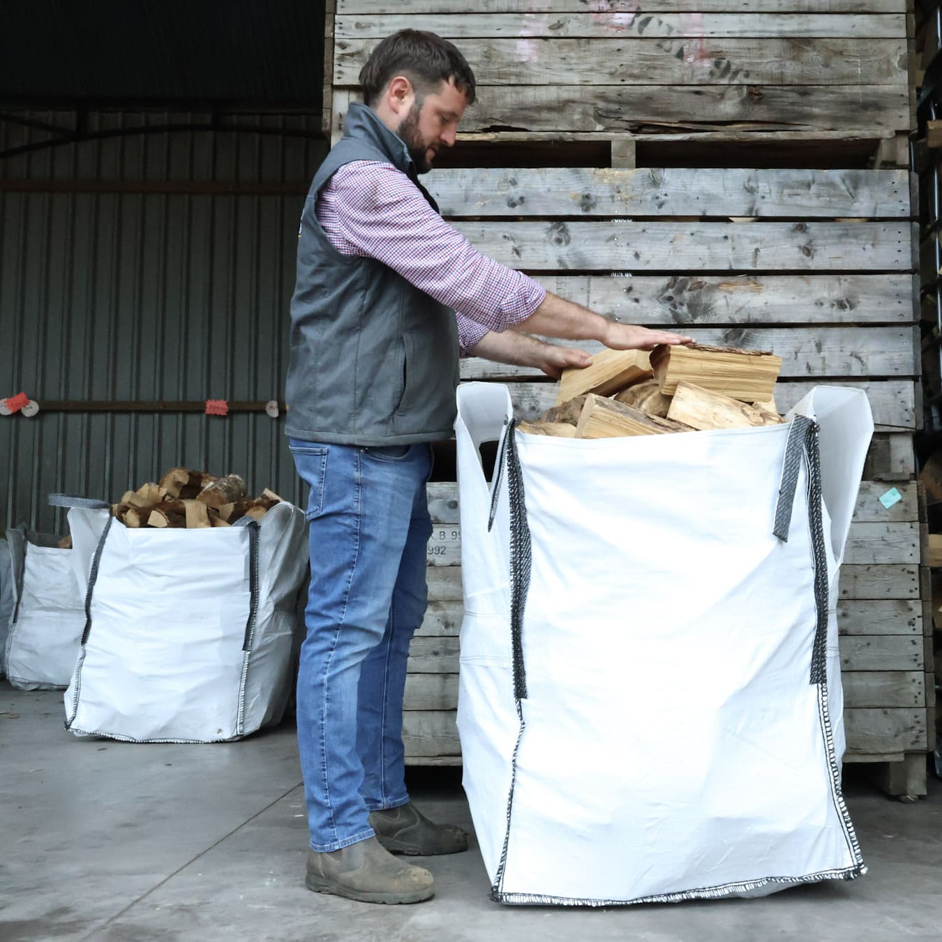 Man loading wood into a large white bag with more bags and wood stacked in the background.