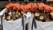 Firewood stacked in a large bag with orange kindling bags on top, against a background of more stacked wood.