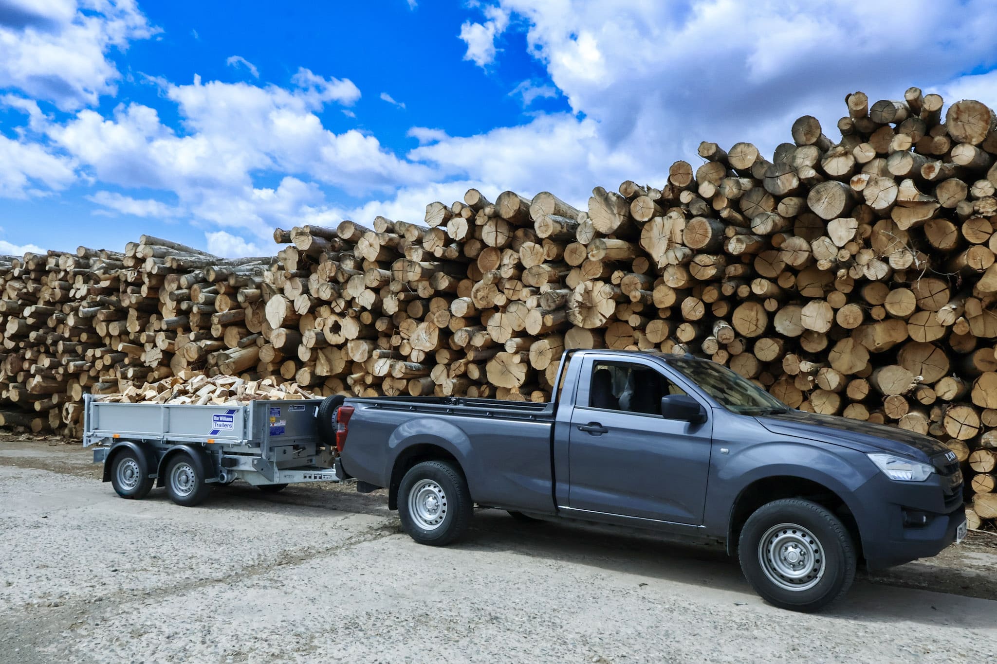 Grey pickup truck with trailer loaded with logs against a blue sky and log stack