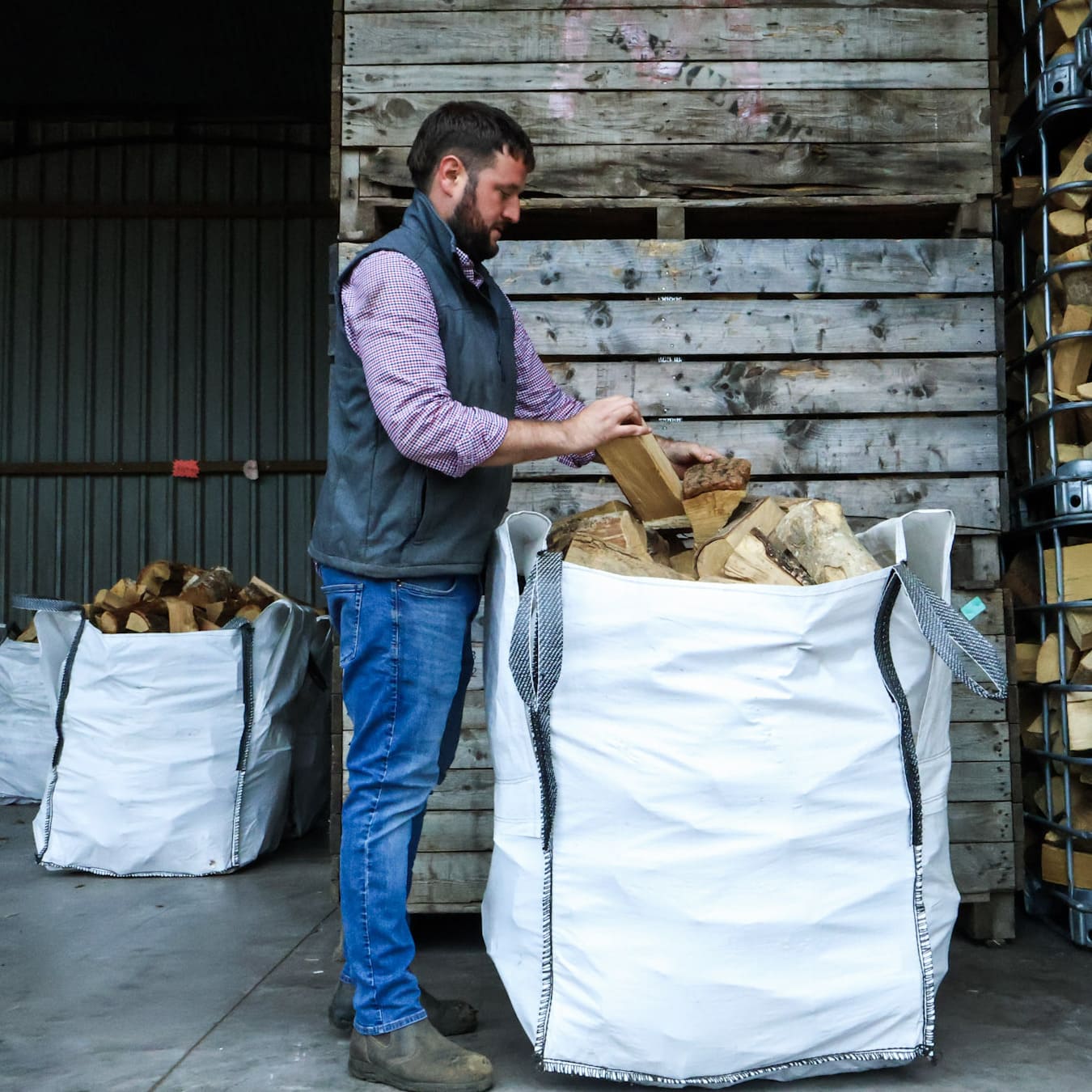 Man loading firewood into a large white bag in front of a wooden wall.